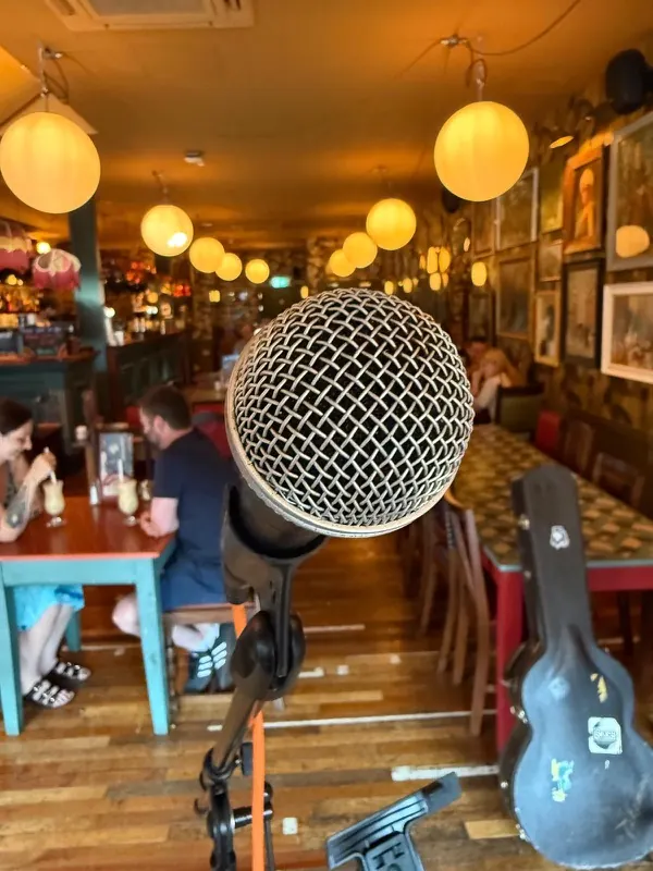 A microphone on its stand at Pedro Lounge, with the warm lamplit room and a guitar case behind