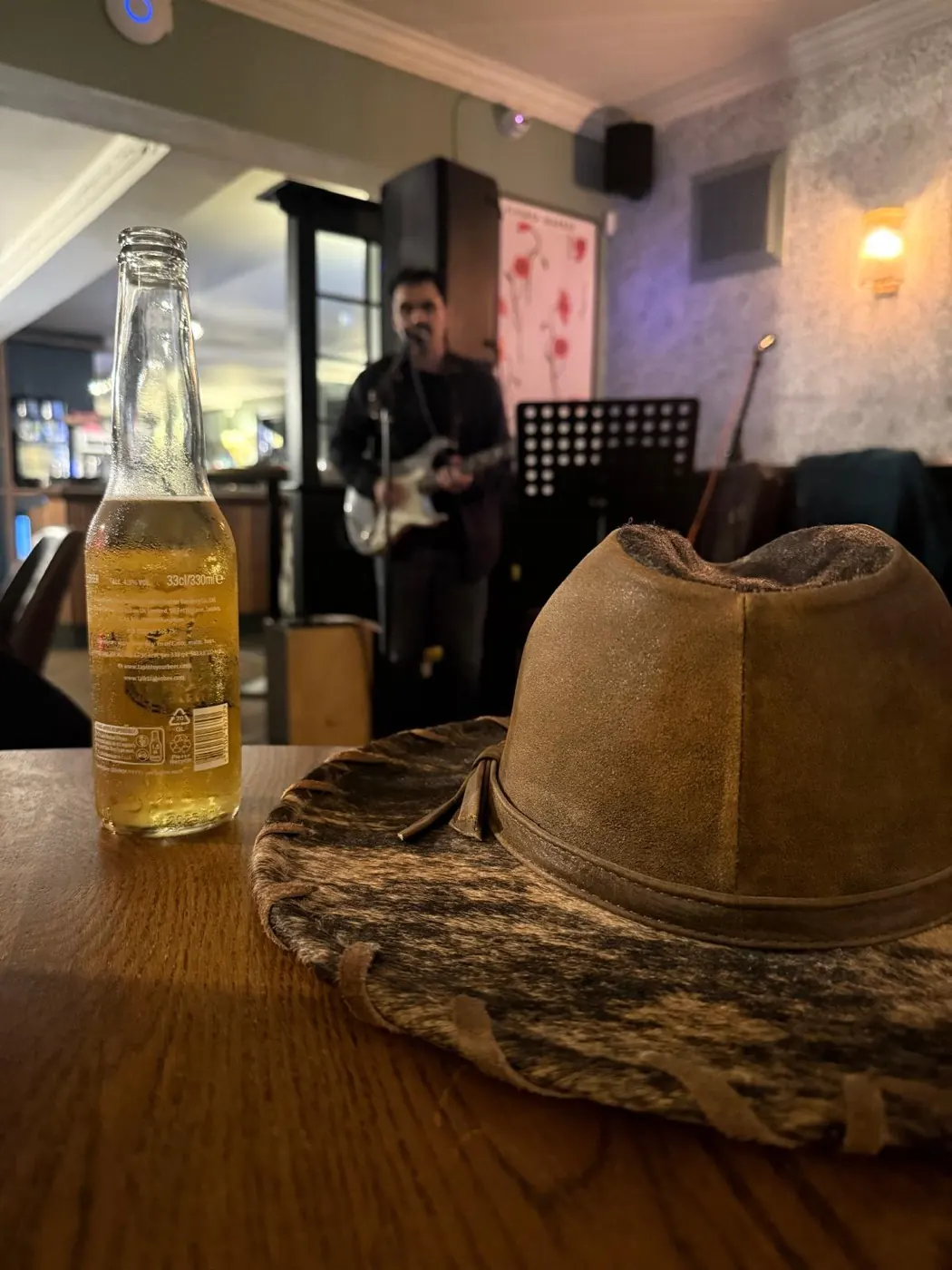 A hat and a bottle of beer on a table at The Oak, with a guitarist singing into the microphone behind
