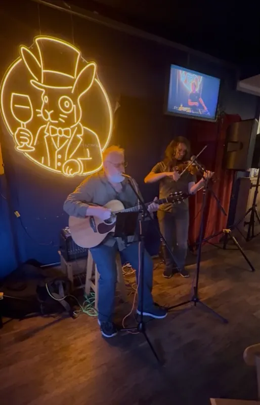 A guitarist and fiddle player performing under the neon rabbit sign at The Rabbit Hole